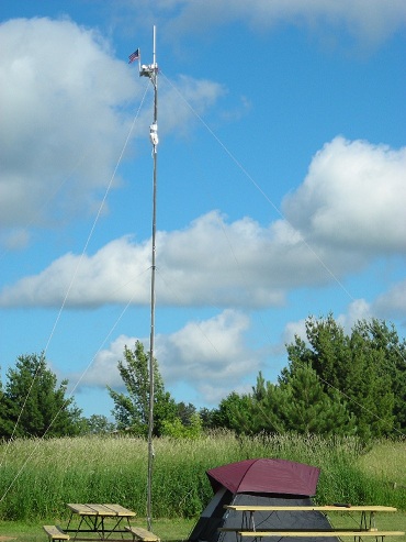 Vertical antenna with small American flag on top at Field Day site with tent and picnic table in foreground.
