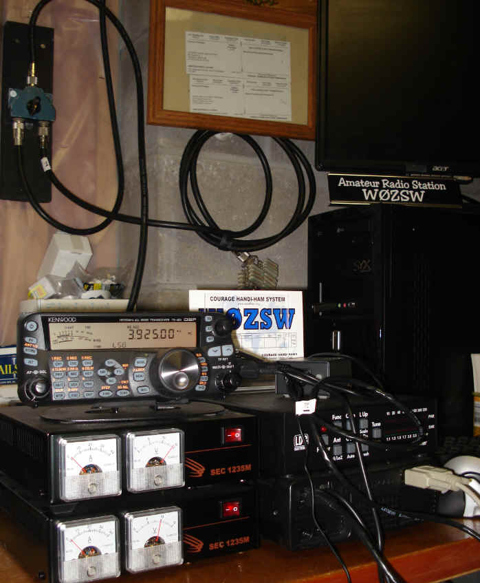 Kenwood TS-480HX with matching SEC switching power supplies, LDG AT-200PRO tuner, RigBlaster interface, rig  control computer, and manual antenna switch mounted on the wall.  